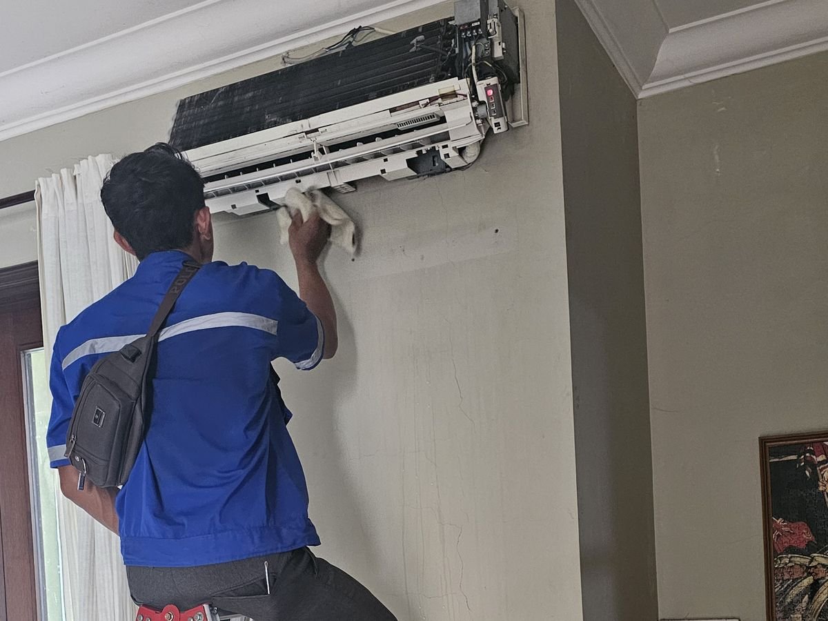 Teknisi di atas tangga membersihkan unit indoor AC yang penutupnya sudah dibuka